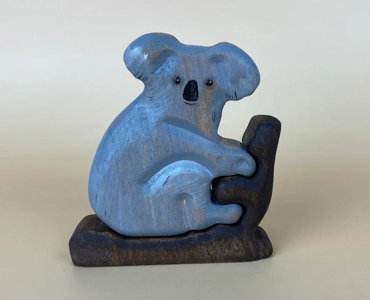 Smiling wooden koala bear toy with blue-grey shade and climbing a branch