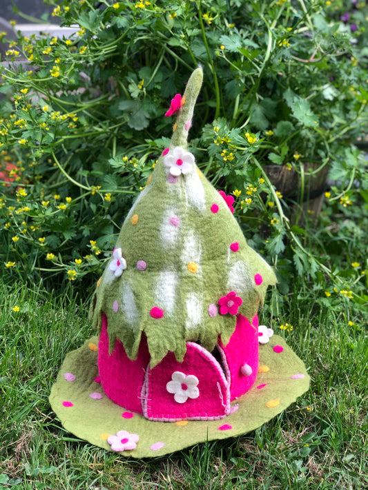 Pink and green felted toy fairy home with sewn on flowers and pointy roof, sitting on a grass patch.